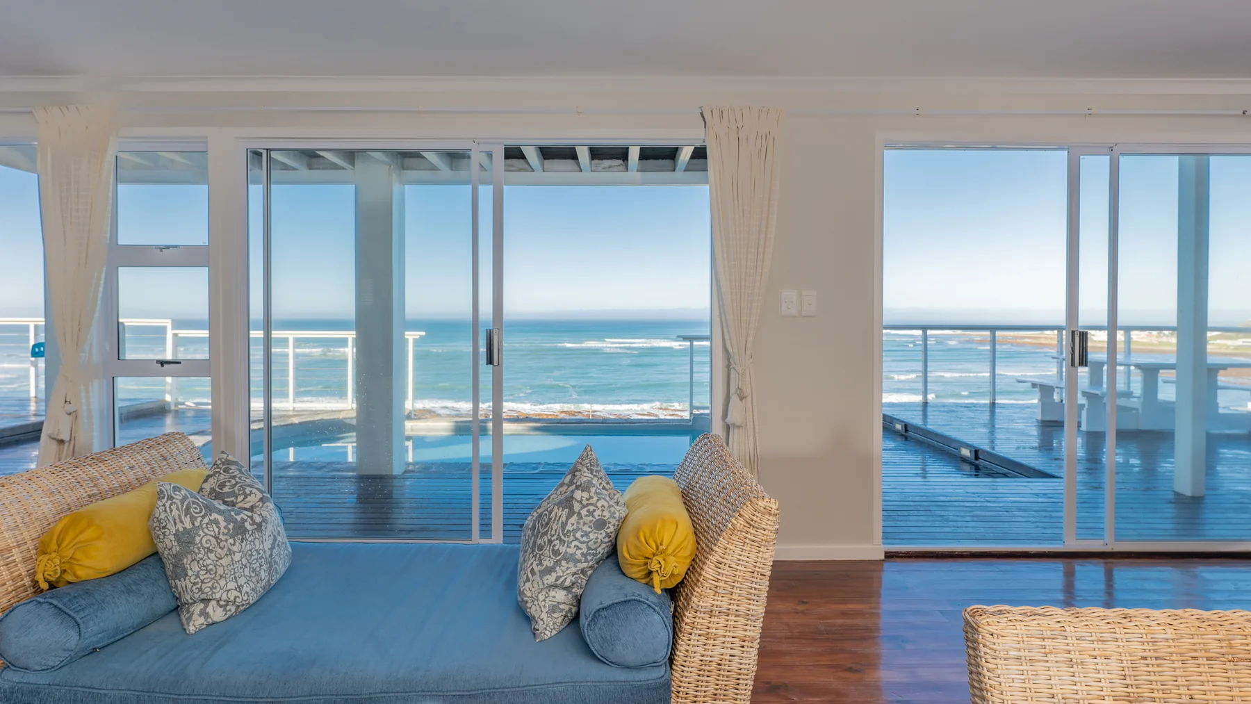 The bright sea-facing sitting room at Driftwood, with wide glass doors opening towards a deck, pool and ocean