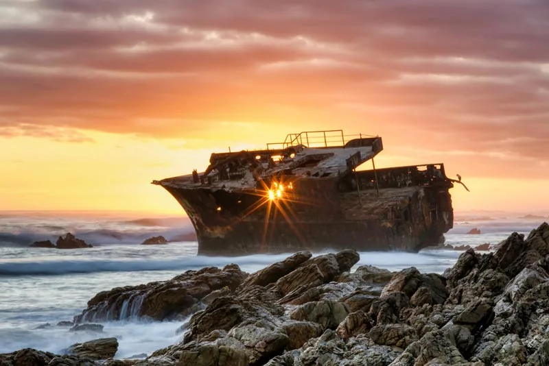 A historic shipwreck silhouetted against a golden sunrise and breaking waves near Cape Agulhas
